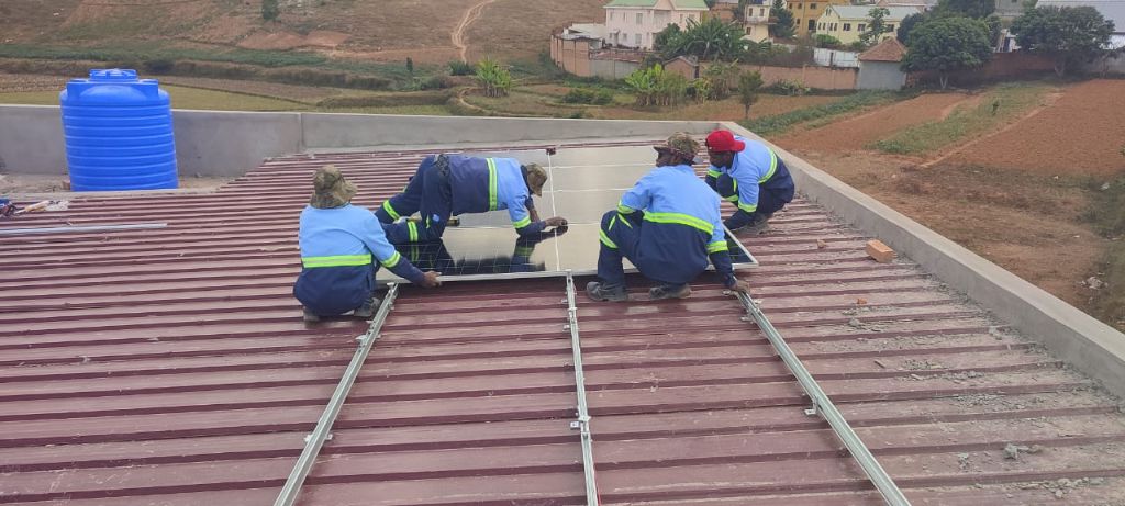 Quatre techniciens Alona en uniformes bleus réglementaires posant un panneau solaire sur toiture rouge — paysage malgache